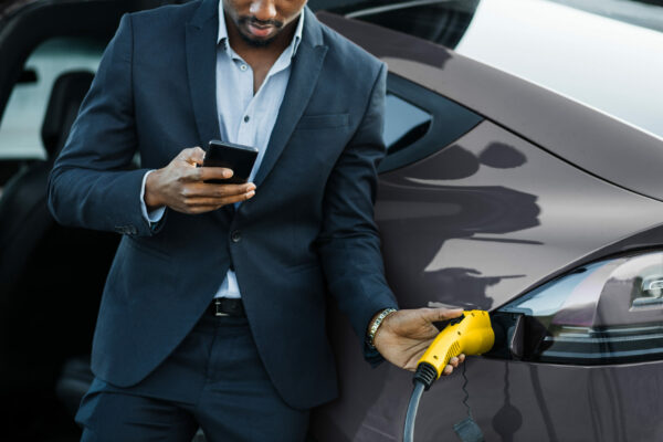 Close up of african businessman unplugging charging cable from electric car while using modern smartphone. Concept of lifestyles, ecology vehicle and busy people.